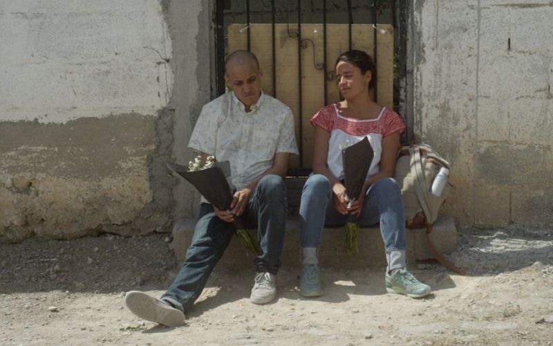 Fotograma de la película Fauna de Nicolás Pereda, con dos personas sentadas afuera de una casa sosteniendo flores.