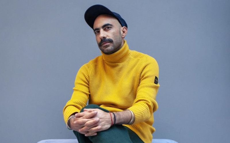 Fernando Frías de la Parra, director del documental Depeche Mode: M, posando con suéter amarillo y gorra frente a un fondo gris.