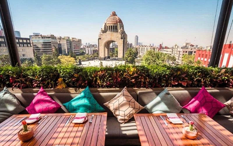 Terraza Cha Cha Chá con vista al Monumento a la Revolución en CDMX, mesas de madera y cojines de colores vibrantes.