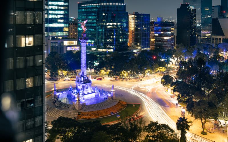 Vista nocturna del Ángel de la Independencia en Paseo de la Reforma, rodeado de edificios iluminados en la CDMX.