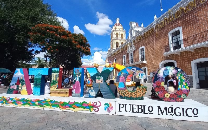 Letras coloridas de Atlixco Pueblo Mágico frente a edificios coloniales y un cielo azul despejado.