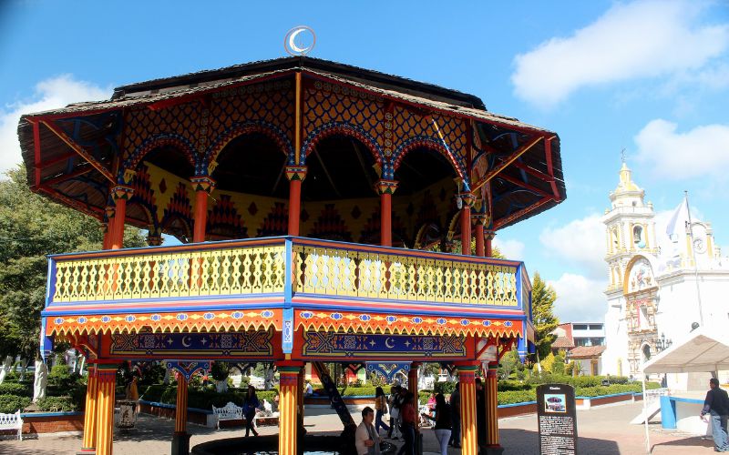 Kiosco colorido de estilo mudéjar en la plaza principal de Chignahuapan con la iglesia del fondo bajo un cielo azul.