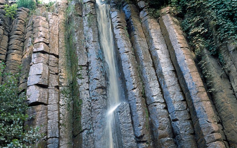Cascada entre formaciones rocosas de los Prismas Basálticos en Huasca de Ocampo rodeada de vegetación.