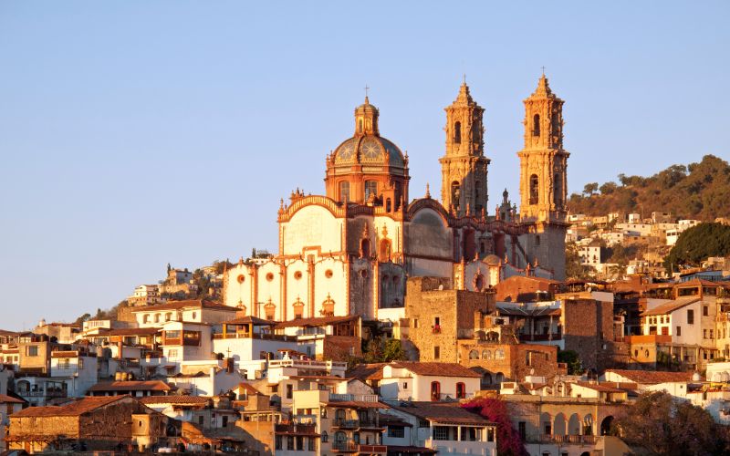 Vista panorámica de Taxco con la Parroquia de Santa Prisca iluminada por el atardecer entre casas coloniales.