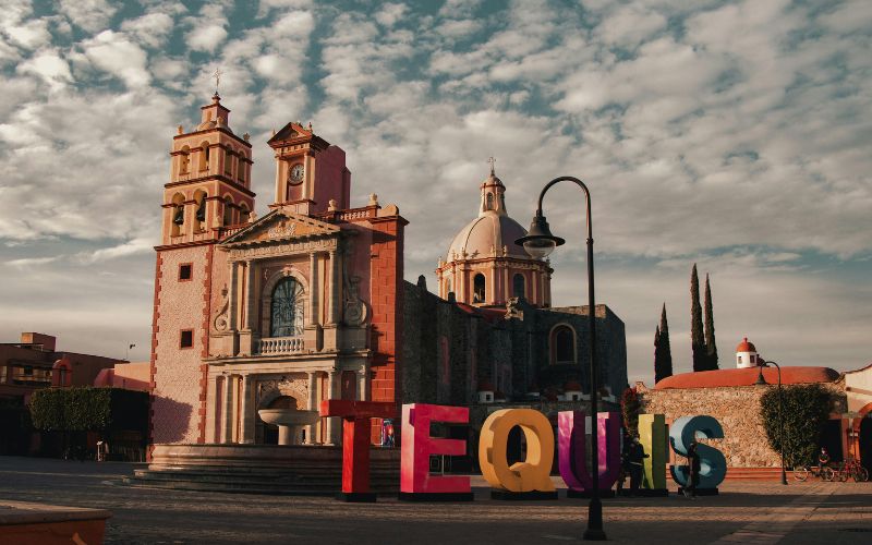 Letras coloridas de Tequisquiapan frente al templo de Santa María de la Asunción bajo un cielo parcialmente nublado.