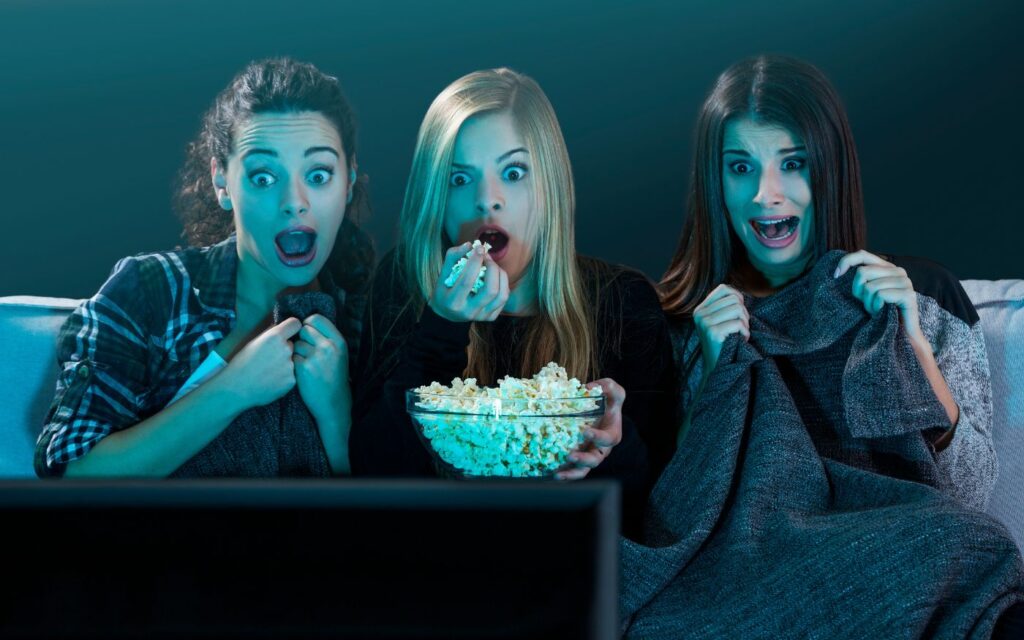 Tres amigas viendo películas de terror mexicano clásico, sorprendidas mientras comen palomitas en la oscuridad.