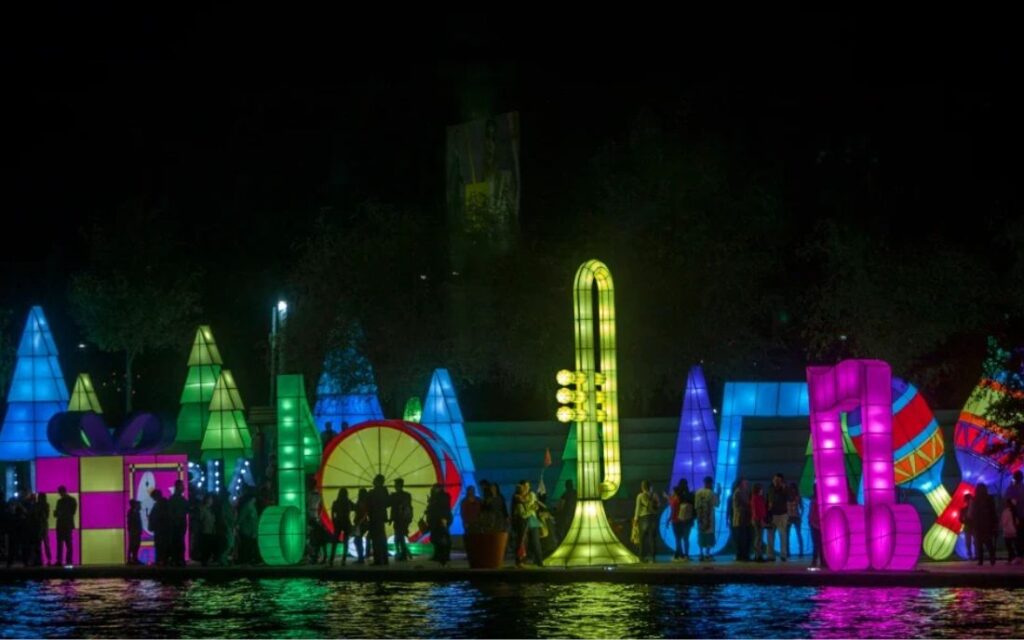 Recorrido nocturno de Luztopía en Monterrey con figuras iluminadas de colores, visitantes caminando y reflejos de luz sobre el agua.
