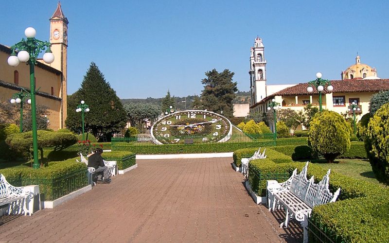 Reloj floral de Zacatlán de las Manzanas en el centro histórico, rodeado de jardines, bancas y edificios coloniales de Puebla.